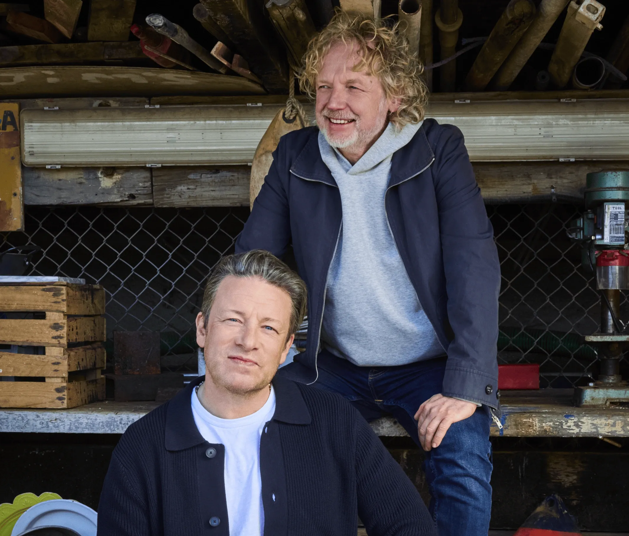 Jamie Oliver and a man with curly hair pose smiling in a rustic workshop.